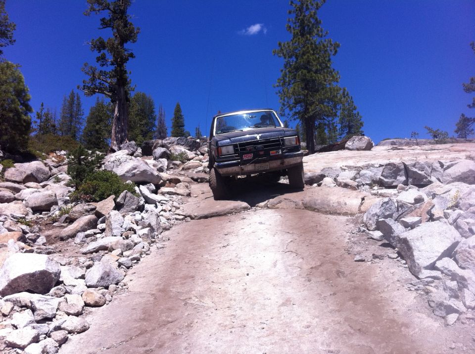 1990 Ford Bronco navigating a rough, rocky off-road path with trees and blue sky in the background