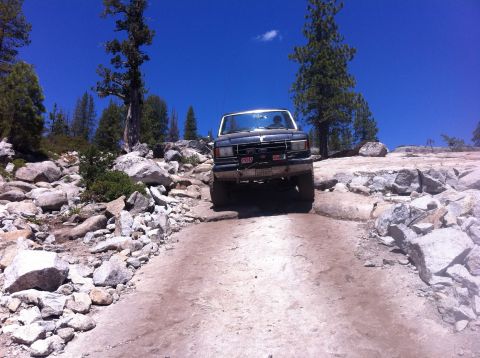 1990 Ford Bronco - This SUV navigates a challenging off-road path under a clear blue sky.