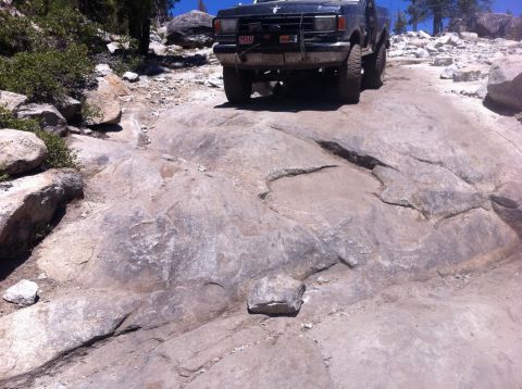 1990 Ford Bronco - A Ford F-Series truck navigates a challenging rocky off-road path under a clear blue sky.