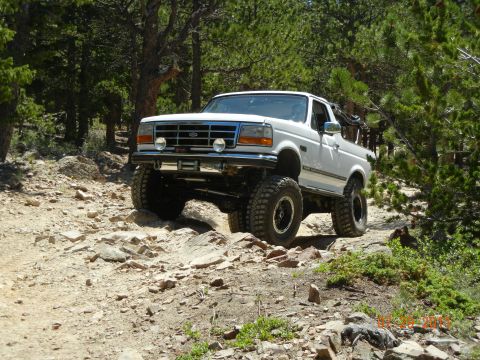 1995 Ford Bronco - A lifted white Ford Bronco with its top removed crawls over a rocky ledge on a pine-forest trail, flexing its suspension with a front tire…
