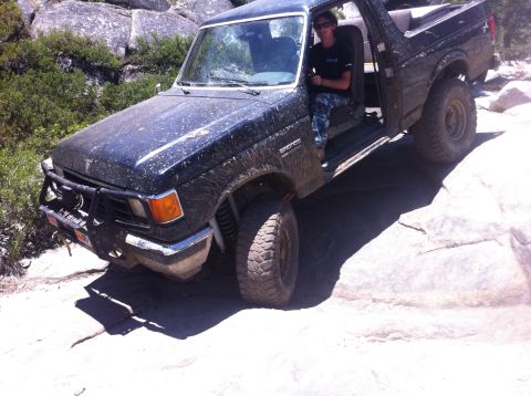 1990 Ford Bronco - A person drives a dark-colored Ford Bronco over a rocky terrain, with the vehicle covered in dust.