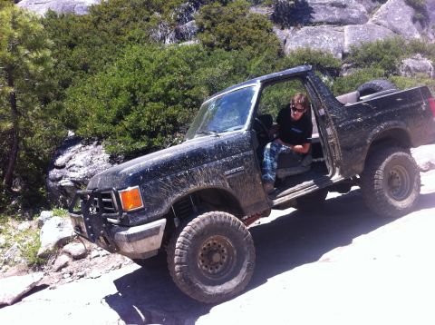 1990 Ford Bronco - A person sits in a muddy Ford Bronco as it navigates a rough, off-road path.