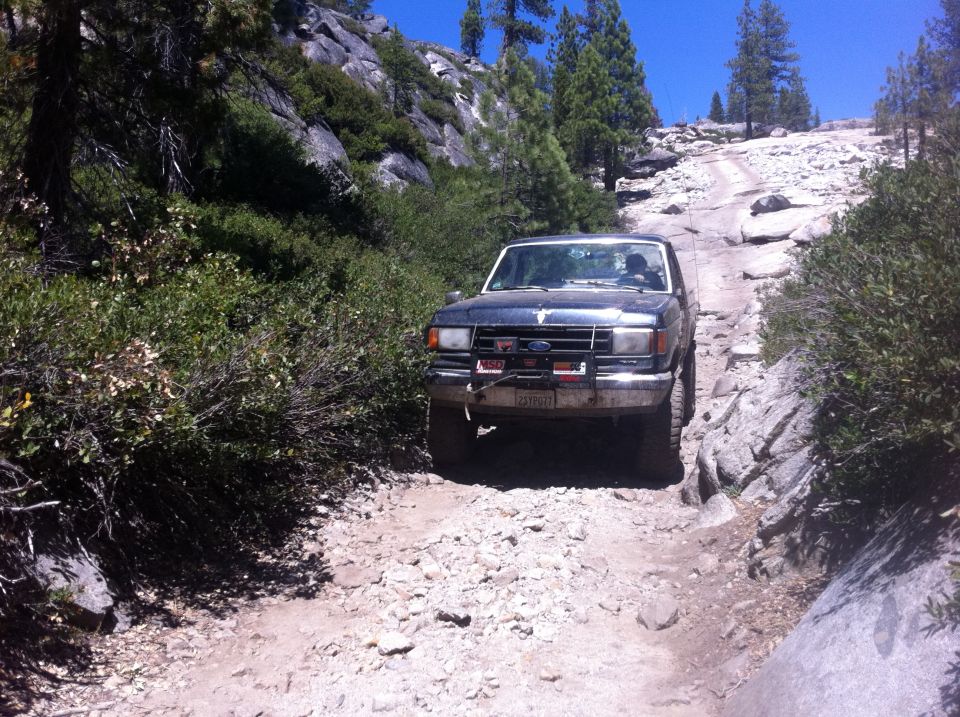 1990 Ford Bronco driving up a steep, rocky, off-road trail with trees and boulders on either side