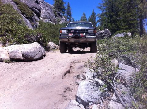 1990 Ford Bronco - A lifted Ford truck navigates a rocky off-road path under a clear blue sky.