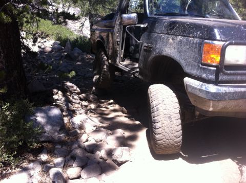 1990 Ford Bronco - A black Ford Bronco navigates a challenging rocky trail with its driver's door ajar.