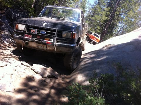 1990 Ford Bronco - A black Ford Bronco navigates a challenging rocky trail, with another orange vehicle visible in the background.