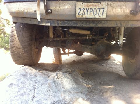 1990 Ford Bronco - The underside of a vehicle navigates rocky terrain, showing its rear axle and suspension system.