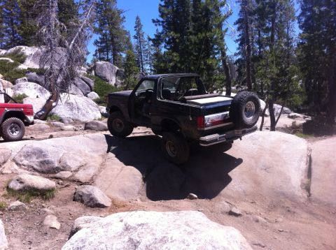 1990 Ford Bronco - A black 1990 Ford Bronco navigates a rocky trail, with another red SUV visible in the background.