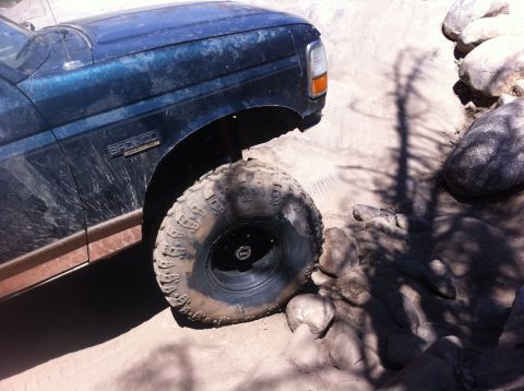 1990 Ford Bronco - A blue Ford Bronco is shown off-roading on a sunny day, with its front tire close to large rocks.