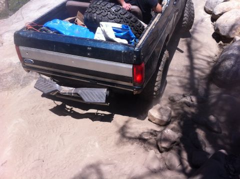 1990 Ford Bronco - Ford pickup truck driving over rocks, with a spare tire and gear loaded in the bed.
