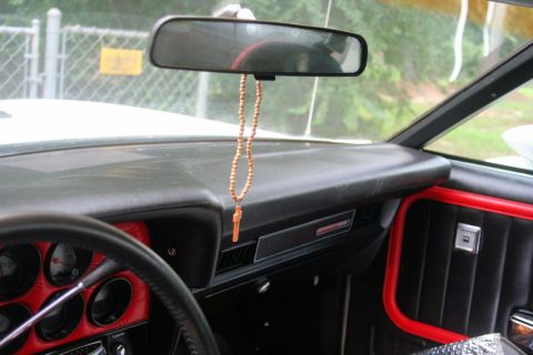 1976 Ford Ranchero - The interior of a 1976 Ford Ranchero shows black upholstery with red accents and a rosary hanging from the mirror.