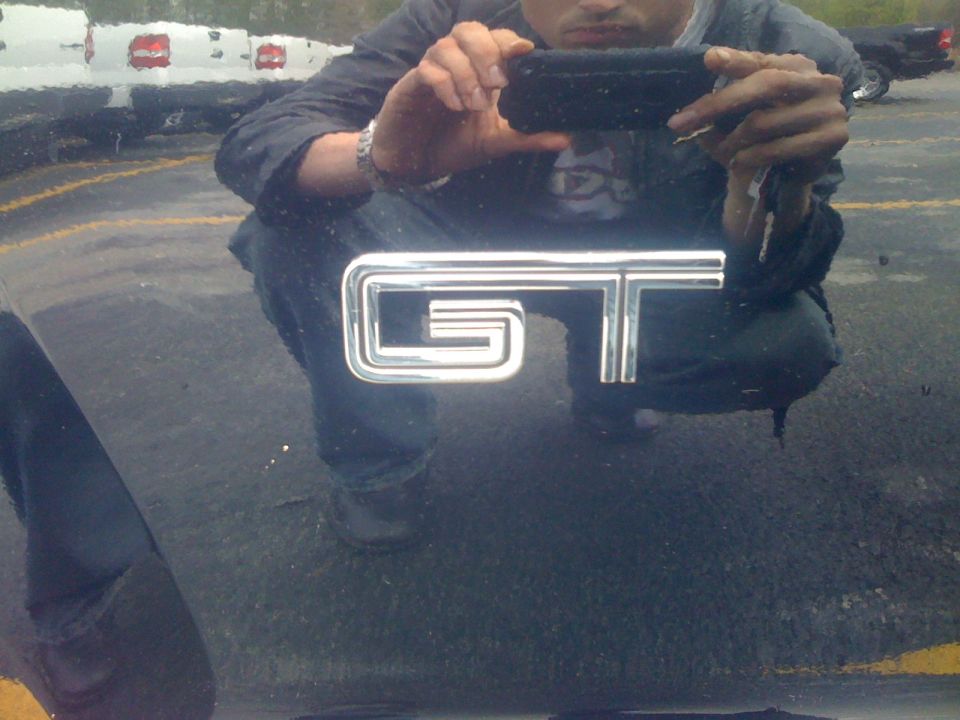 Black car hood with chrome GT emblem, reflection of person holding phone, other vehicles in background