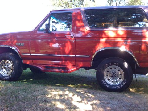 1996 Ford Bronco - This red Ford Bronco SUV is parked on a grassy area with trees casting shadows.