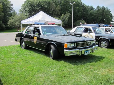 1991 Ford LTD Crown Victoria - black-and-white Chevrolet Caprice police cruiser marked 154 with roof light bar parked on grass under a canopy tent.