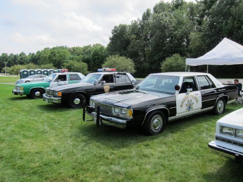 1991 Ford LTD Crown Victoria - lineup of vintage police cruisers on a grassy field, led by a black-and-white highway patrol sedan with star emblem.