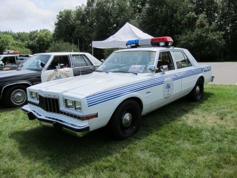 1991 Ford LTD Crown Victoria - white Plymouth police sedan with blue accent stripes, a circular seal and POLICE lettering parked on grass under a tent.