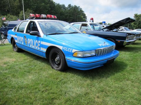 1991 Ford LTD Crown Victoria - blue-and-white NYPD Chevrolet Caprice police cruiser with roof light bar parked on grass at a car show.