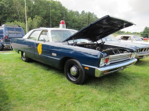 1991 Ford LTD Crown Victoria - dark blue vintage Plymouth police sedan with a yellow door shield and raised hood parked on grass at a show.