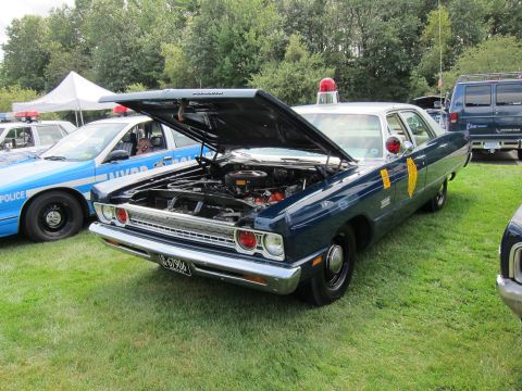 1991 Ford LTD Crown Victoria - dark blue vintage Plymouth police sedan with its hood raised to show the engine, parked beside a blue NYPD cruiser on grass.