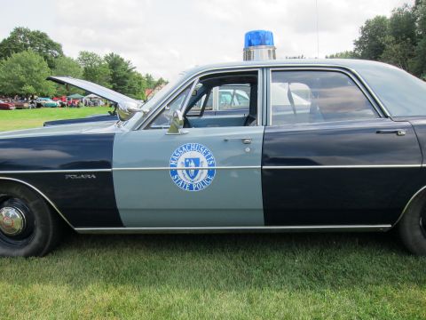 1991 Ford LTD Crown Victoria - Side profile of a two-tone blue Plymouth Polara bearing a Massachusetts State Police seal on the door, parked on grass.