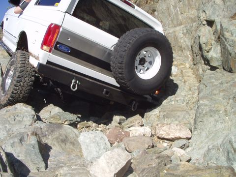 1993 Ford Bronco - White Ford Bronco navigates a rocky terrain, showing its rear end and spare tire.
