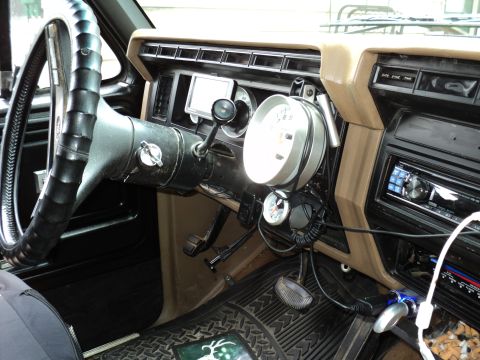 1986 Ford Bronco - The dashboard of an older truck features a large steering wheel and several aftermarket gauges, including a prominent tachometer.