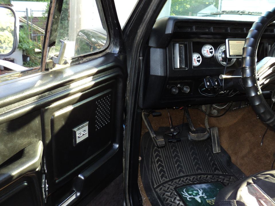 Interior view of a 1980s Ford truck showing door panel, dashboard with gauges, steering wheel, and pedals