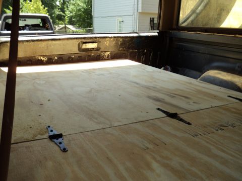1986 Ford Bronco - The interior of a truck bed is shown with a newly installed plywood floor. Another vehicle is visible through the rear window.