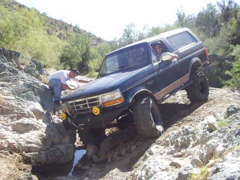 1993 Ford Bronco - A dark green Ford Bronco is driven over a rocky, off-road course with a person assisting.