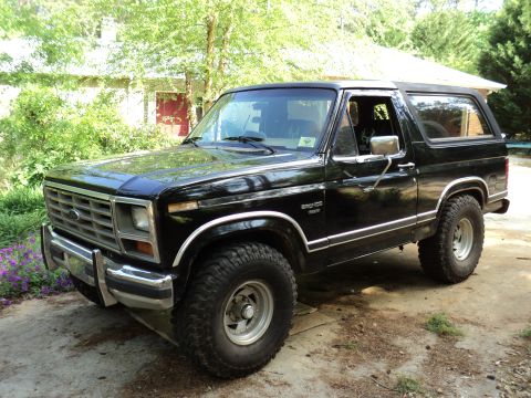 1986 Ford Bronco - This 1986 Ford Bronco is shown in a three-quarter front view, parked in a driveway with trees and a house in the background.