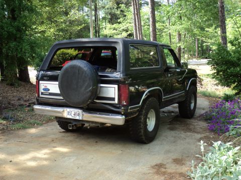1986 Ford Bronco - This black Ford Bronco is parked outdoors on a concrete driveway, surrounded by trees and foliage.