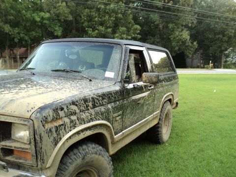 1986 Ford Bronco - A dirty black SUV sits on a grassy field, its body covered in mud.