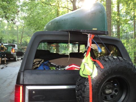 1986 Ford Bronco - An SUV loaded with a canoe and gear is parked near other vehicles in a wooded area.