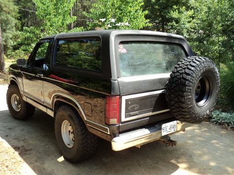 1986 Ford Bronco - This black Ford Bronco is parked outdoors with trees in the background. It has a spare tire mounted on the rear tailgate.