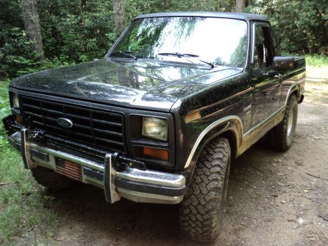 1986 Ford Bronco - This black Ford Bronco is parked on a dirt path surrounded by trees.