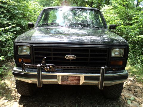 1986 Ford Bronco - Front view of a 1986 Ford Bronco, equipped with a winch and an American flag decal on the bumper, parked on a dirt road.