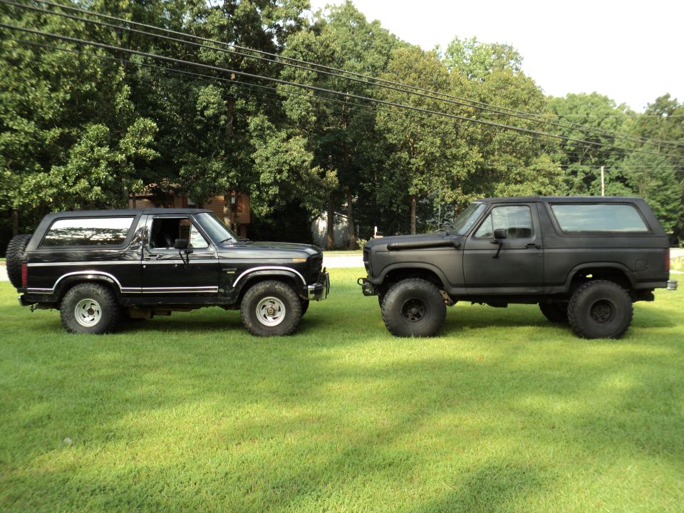 Two Ford Broncos parked on grass, one black, one matte black. The black Bronco is a 1980s model