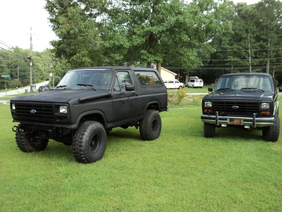 Two matte black Ford Broncos parked on a grassy lawn. The Bronco on the left is lifted with large tires