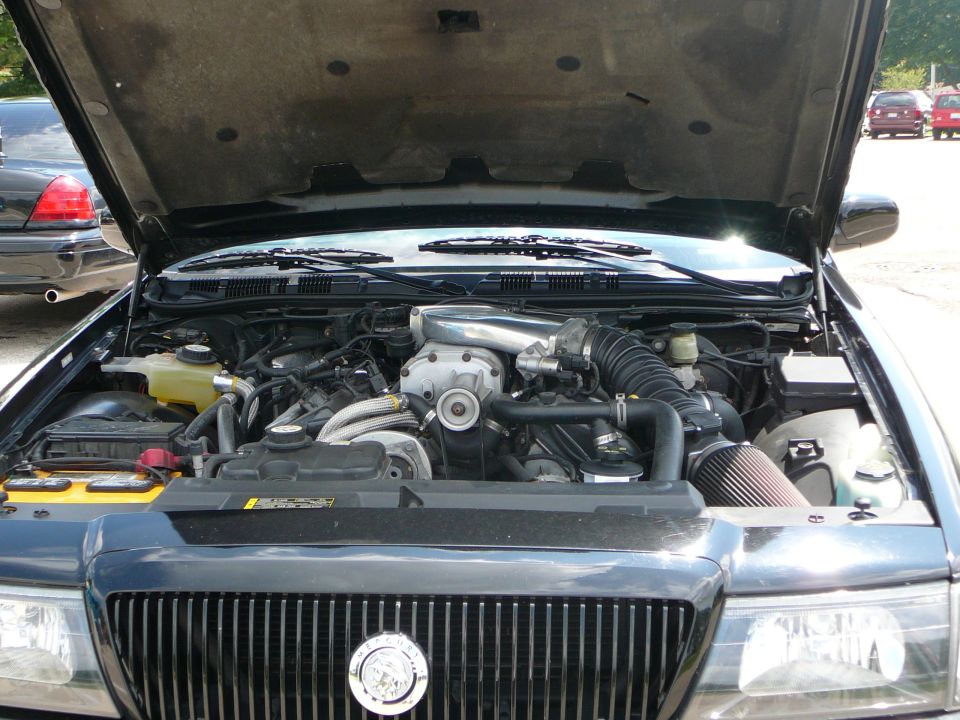 1999 Mercury Grand Marquis engine bay with supercharger and aftermarket air filter. Another car is visible in the background