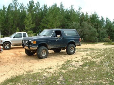 1995 Ford Bronco - A lifted Ford Bronco and a pickup truck are parked off-road near a wooded area.