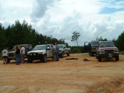 1995 Ford Bronco - A group of off-road vehicles are gathered on a dirt lot under a cloudy sky.