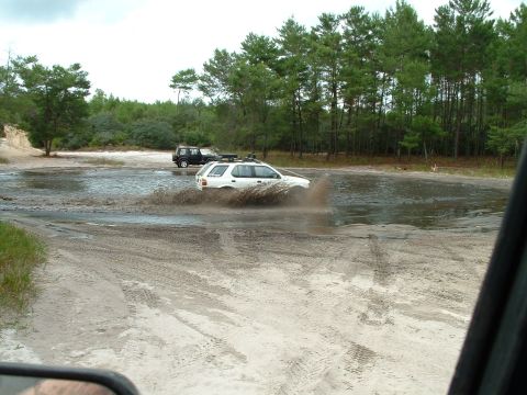 1995 Ford Bronco - Two SUVs navigate a muddy water crossing with trees and sand in the background.