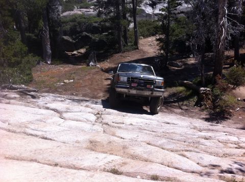 1990 Ford Bronco - A gray pickup truck navigates a rough, rocky path through a wooded area. The Supermotors.net logo is visible in the lower right corner.
