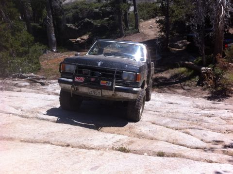 1990 Ford Bronco - A black Ford Ranger navigates a challenging off-road path with trees and rocks surrounding it.