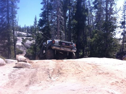 1990 Ford Bronco - A dark green 1987 Ford Bronco navigates a challenging off-road trail in a wooded, rocky area.