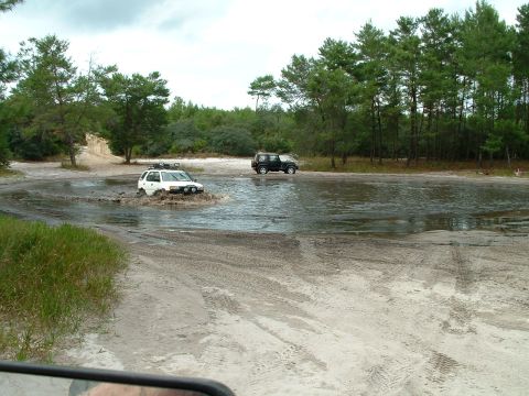 1995 Ford Bronco - Two SUVs navigate a muddy water crossing on a dirt path surrounded by trees.