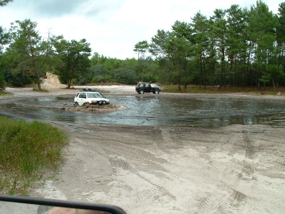 Two SUVs driving through a muddy puddle on a dirt road with trees in the background