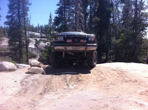 1990 Ford Bronco - Modified black SUV navigates a rocky, off-road trail with tall trees in the background.