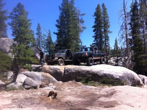 1990 Ford Bronco - Two pickup trucks are parked on large rocks in a wooded, mountainous area under a clear blue sky.