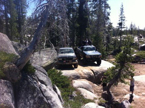 1990 Ford Bronco - Two trucks traverse a rocky, wooded landscape, with several people enjoying the off-road adventure.